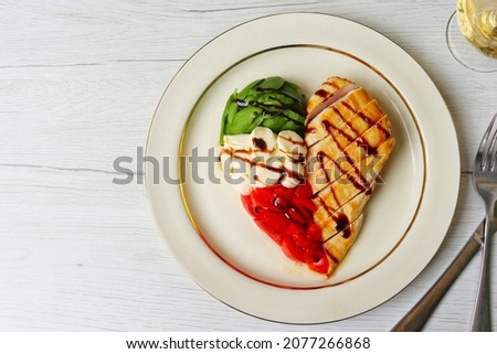 Heart shaped Italian Caprese Salad arranged by Italian basil,buffalo mozzarella and tomatoes look like Italian flag and grilled chicken breast on top with balsamic vinegar on plate with white table