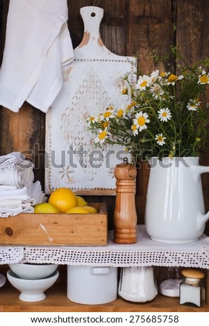 Closeup of two white wood rustic kitchen shelves with assorted household items. Various objects, include, pepper shaker, cups, pitchers bottles, lemons and bouquet of chamomile flowers.
