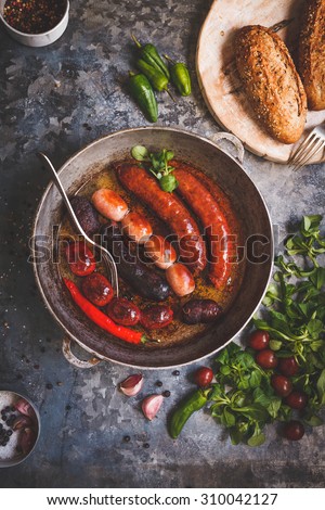A variety smoked colorful sausages served in pan from above on rustic metallic table with bread baguette and veggies. Rustic dark styling. Spanish barbecue tapas.