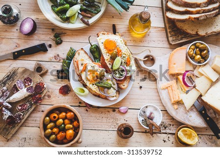 A Toasted sandwich with smoked salmon and fried eggs and variety cheese cutting board with bowl olives and green leaves salad from above a wooden table. Country simple mediterranean food.