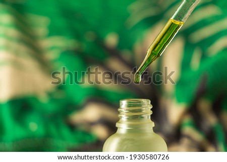 Dropping essential oil into glass bottle on blurred green background.  Detail of a CBD oil dropper against a background of blurry green leaves. Cosmetic oil drips off the pipette.