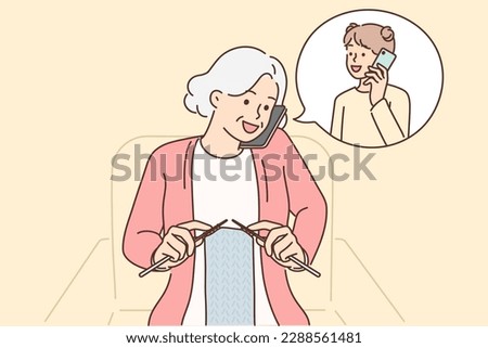 Grandmother makes phone call to granddaughter and knits scarf sitting in chair. Elderly woman with phone is talking to granddaughter, asking how things are at school or inviting to come visit 
