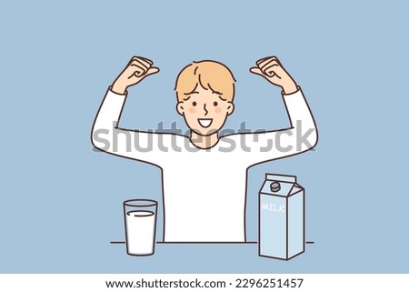 Teenage boy drinks cows milk and flaunts muscles showing off strength gained from eating lactose. Child stands near table with package of milk useful for child organism and immunity