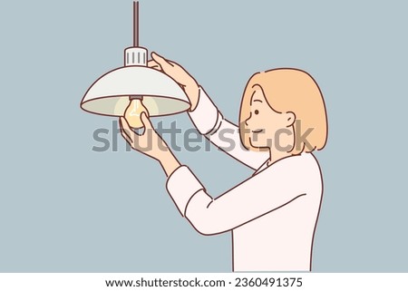 Woman independently changes burned-out light bulb in order to achieve high-quality lighting in house. Girl with smile inserts light bulb into chandelier hanging under ceiling, without help electrician