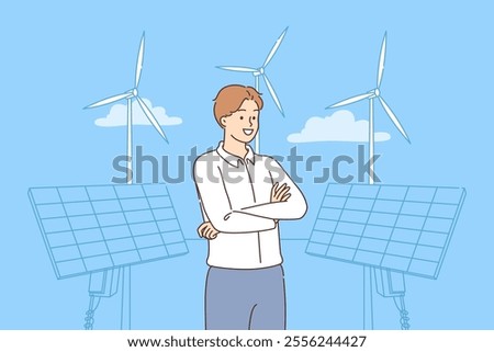 Man stands near ecological power plant with solar panels and wind turbines under blue sky. Businessman owner of regenerative power plant introduces green technology to produce fuel and electricity