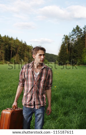 Casual young man with luggage Casual young man standing in green grass in the countryside with his luggage while traveling on vacation