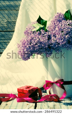 Lilac bouquet in the jug decorated with a pink bow with a gift red box on a wooden surface in vintage style. Vintage Bouquet of lilac flowers