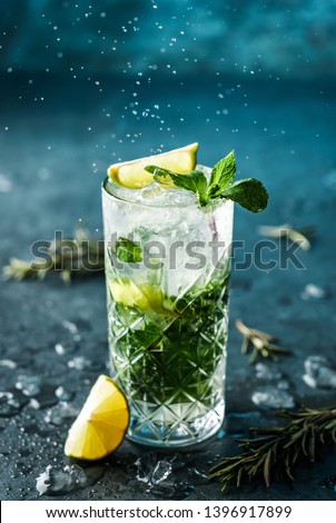 Fresh Mojito cocktail with lime, rosemary, mint and ice in jar glass on dark blue background. Studio shot of drink in freeze motion, drops in liquid splash. Summer cold drink and cocktail