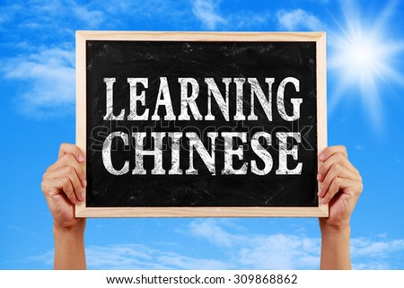 Hands holding blackboard with text Learning Chinese against blue sky background.