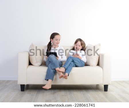 Two young girls sitting on a couch using modern electrical devices.