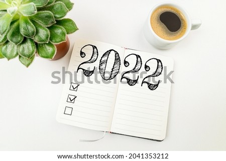New year goals 2022 on desk. 2022 resolutions list with notebook, coffee cup on white desk. Resolutions, plan, goals, action, checklist, idea concept. New Year 2022 