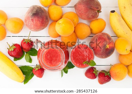 Healthy strawberry banana smoothie with mint in a glass and pitcher on white wooden background. Fresh fruits bananas, peaches and apricots background. Selective focus. Top view