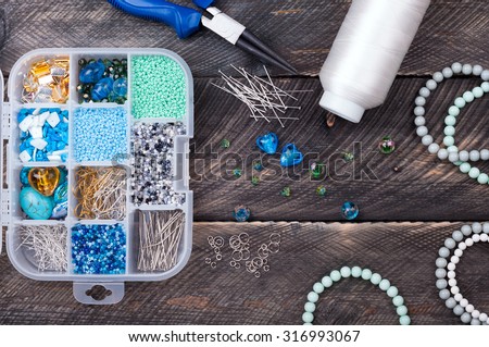 Box with beads and pins, spool of thread, plier and glass hearts to create hand made jewelry on old wooden background. Handmade accessories. Top view