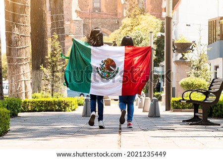 two young mexican women running on a walkway on a tree-lined street, with the mexican flag