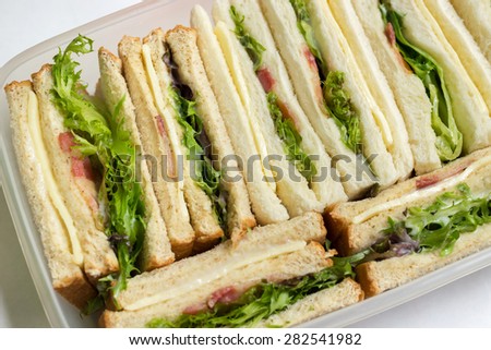 Sandwiches in box for a picnic ; whole wheat and white bread, baby green and red oak leaf lettuce, bacon, cheese and mayonnaise