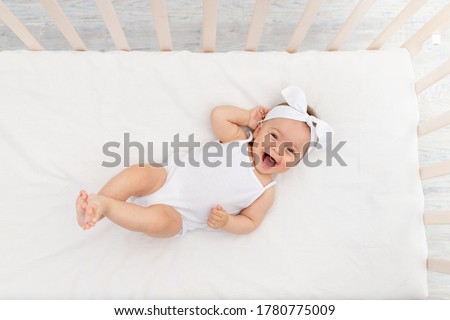 baby girl 6 months old lies in a crib in the nursery with white clothes on her back and laughs, looks at the camera, baby's morning, baby products concept