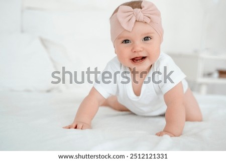 A baby girl is smiling at home on a white bed with a bow on her head, a happy healthy child of six months is laughing.