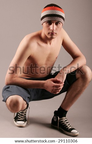 Young man in knitted cap, shorts and gym-shoes sitting on grey background