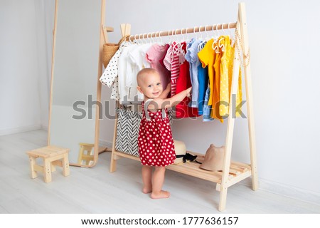 Baby girl stands near a wardrobe and chooses a dress and smiles. Dressing closet with clothes arranged on hangers. Wardrobe of newborn, kids, toddlers, babies full of all clothes. montessori wardrobe.