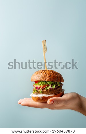 Hand holding burger. Hamburger with meat free plant based cutlet, tomatoes, onions, white sauce and microgreens on blue background. Healthy vegan or vegetarian food concept. Copy space.