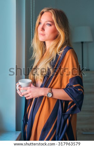 Young sexy woman in underwear holding cup of coffee next to window in the morning
