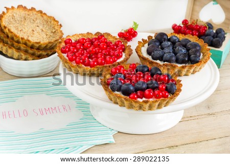 Tartlets with blueberries and red currant on a white cake stand. Empty pie shells and fresh fruits in the background. Colorful decoration.