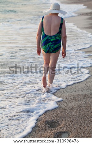 Seventy-old woman in a swimming suit and hat going away along coastline