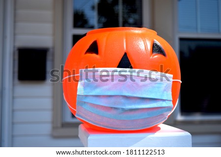 Coronavirus Halloween Pumpkin. Halloween jack o lantern wearing a covid-19 face mask and placed outside a home at daytime. House, front door, and window visible in background.