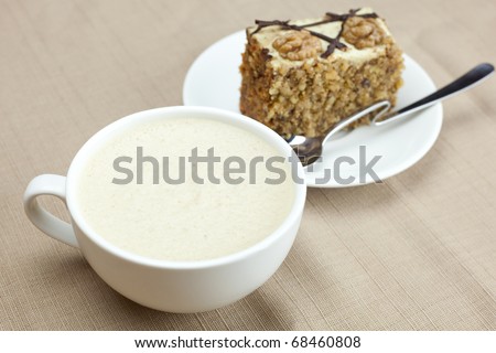 A cup of cappuccino, a piece of cake with nuts and spoon lying on a plate