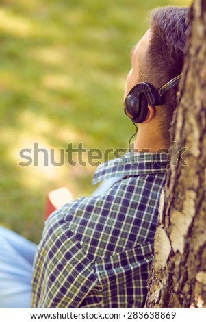 Music, technology, people and leisure concept - close up of teenage boy in headphones with book listening to music online at park