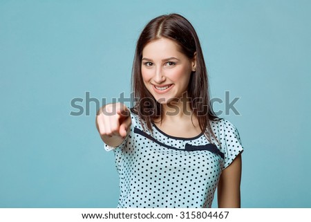 Young brunette woman pointing by a finger at you on a blue background.