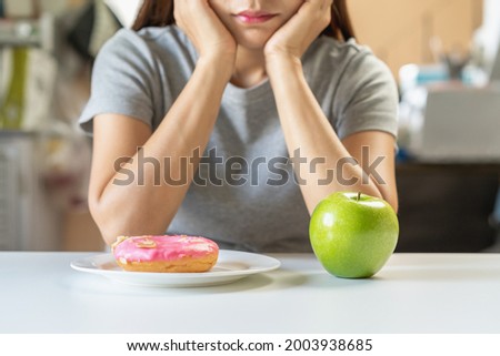 Diet, Dieting asian young woman or girl confused choose, choice green apple or pink sweet donut on table at home, eat food for good healthy, health when hungry. Close up female weight loss person. Foto stock © 