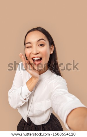 Young woman wearing white shirt with dark long hair standing isolated on bage background taking selfie photo winking to camera posing smiling playful