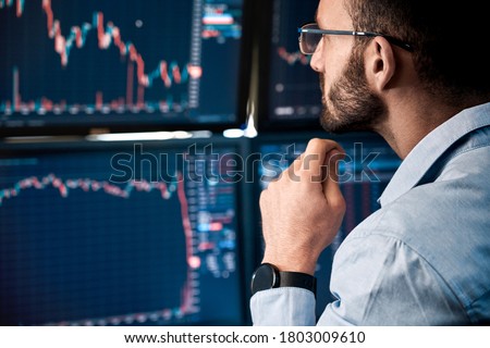 Bearded man trader wearing eyeglasses sitting at desk at office monitoring stock market looking at monitors analyzing candle bar price flow touching chin concerned trading concept close-up