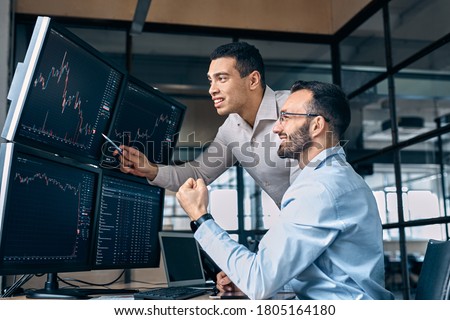 Two men traders sitting at desk at office together monitoring stocks data candle charts on screen analyzing price flow smiling cheerful having profit teamwork concept