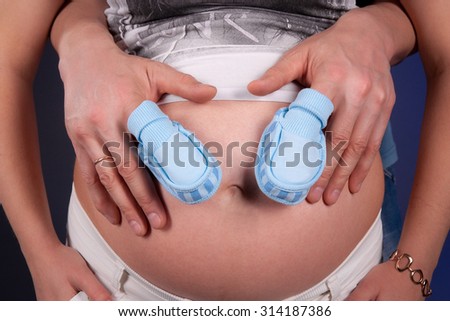 Pregnant belly with fathers hands holding belly and small blue shoes. Close up horizontal photo