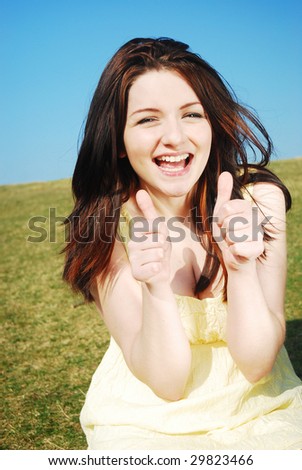 Beautiful young woman giving you a thumbs up and smiling in a field with a blue sky.