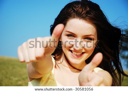 A beautiful young woman giving you a thumbs up and smiling in a field with a blue sky.