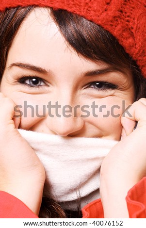 A close up of a woman wearing a hat and scarf, pulling the scarf up over her face, on a white background.