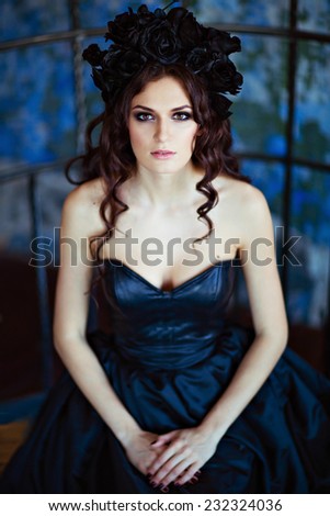 Curly beautiful brunette with a wreath of black flowers sitting on a blue background