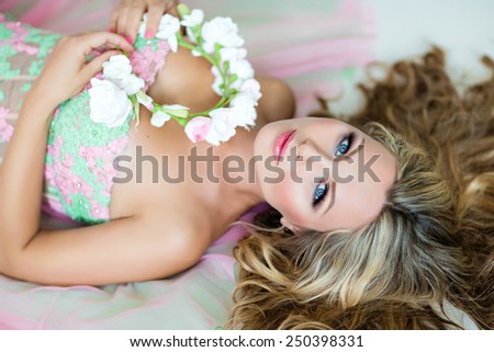 Very beautiful sexy sensual girl with curly blond hair, blue eyes in a lace dress and a wreath of delicate spring flowers, lying on the white floor, close u