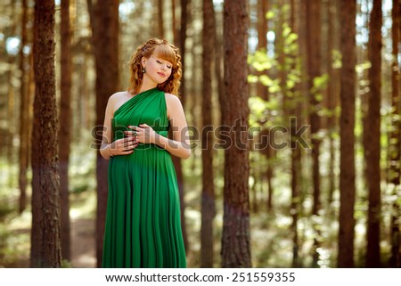 Portrait of a beautiful curly haired pregnant girl with her eyes closed in the green dress in the woods among the pines