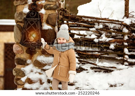 Very sweet beautiful little girl child in a beige coat smiling against winter village near the lantern