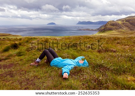 The girl in the blue jacket lying on the grass on a background of the ocean and mountains in Norway