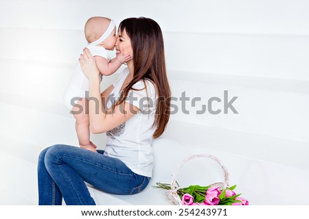 little smiling baby daughter bites his nose mom brunette in white t-shirt and jeans on a white background in the Studio, next to the tulips