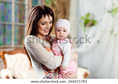 Happy and beautiful mom brunette holding a charming little daughter baby in the interior in beige tones