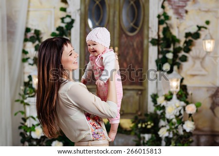 Happy and beautiful mom brunette holding a charming little daughter baby and laughing together, in the interiors of the house in beige