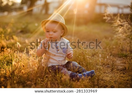 Small charming toddler boy hat sitting in the grass at sunset in the summer and blows on the straw
