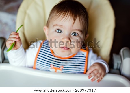 Little adorable baby boy sitting in high chair, holding a spoon in his hands and pulls her lips into a tube