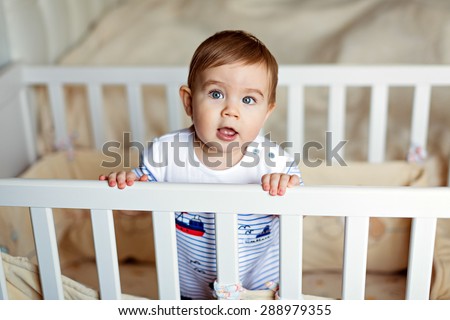 Little cute adorable little blond boy in a striped bodykit is in the nursery with white crib and stares into the camera